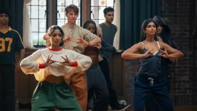 "A diverse group of young adults practicing dance in a studio with wooden floors, brick walls, and large windows. The two women in the front are focused on their hand movements, suggesting a choreographed routine."