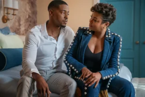 "A man in a white shirt and a woman in a blue embellished outfit sit together on a bed in a cozy, well-lit bedroom, engaged in a serious conversation."